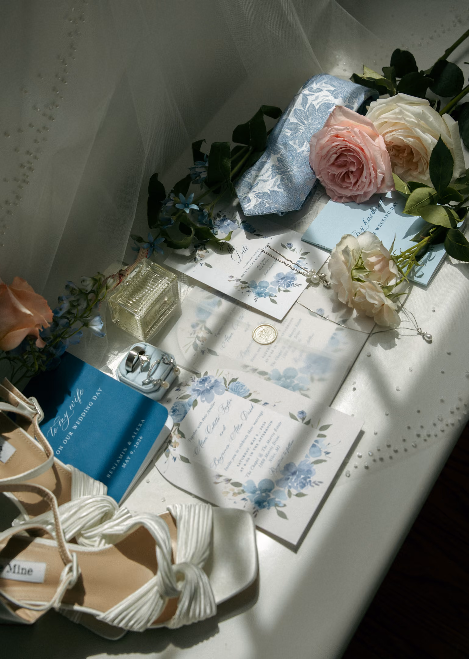 Wedding details flat lay with white shoes, blue floral invitations, rings, and pink and white roses.