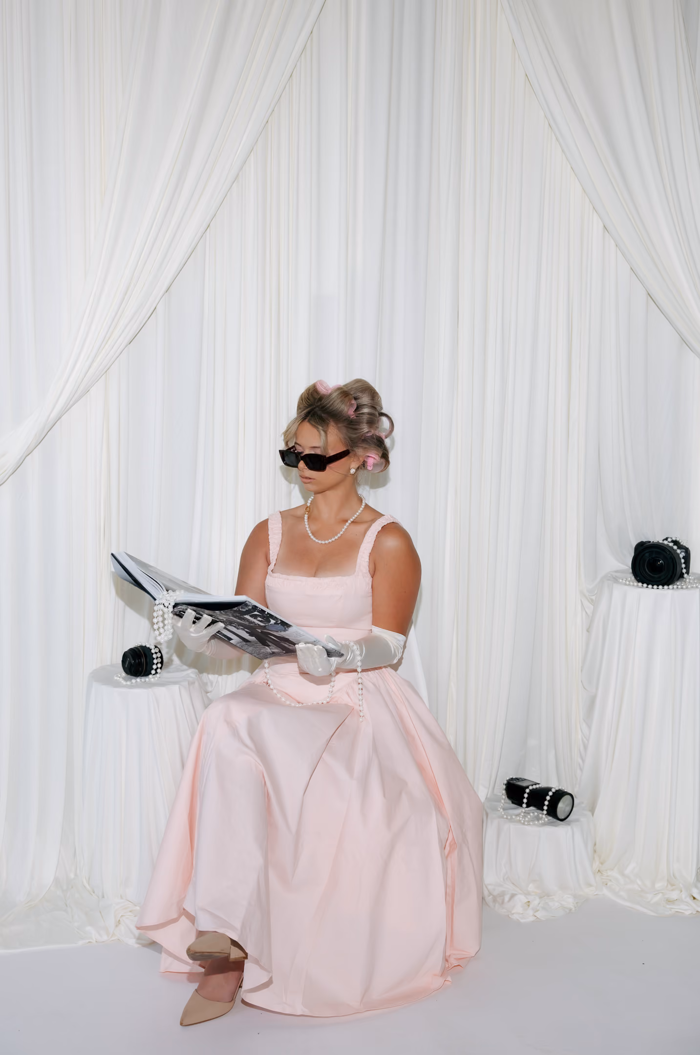 Woman in pink dress with gloves and hair rollers reading a book in a white draped room.