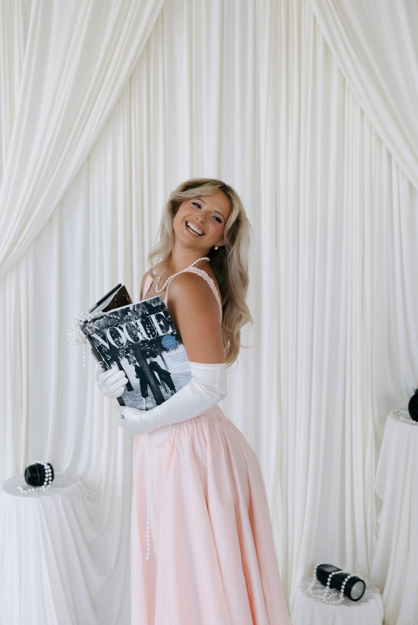 Smiling woman in pink dress and white gloves holding a Vogue magazine against white draped background.