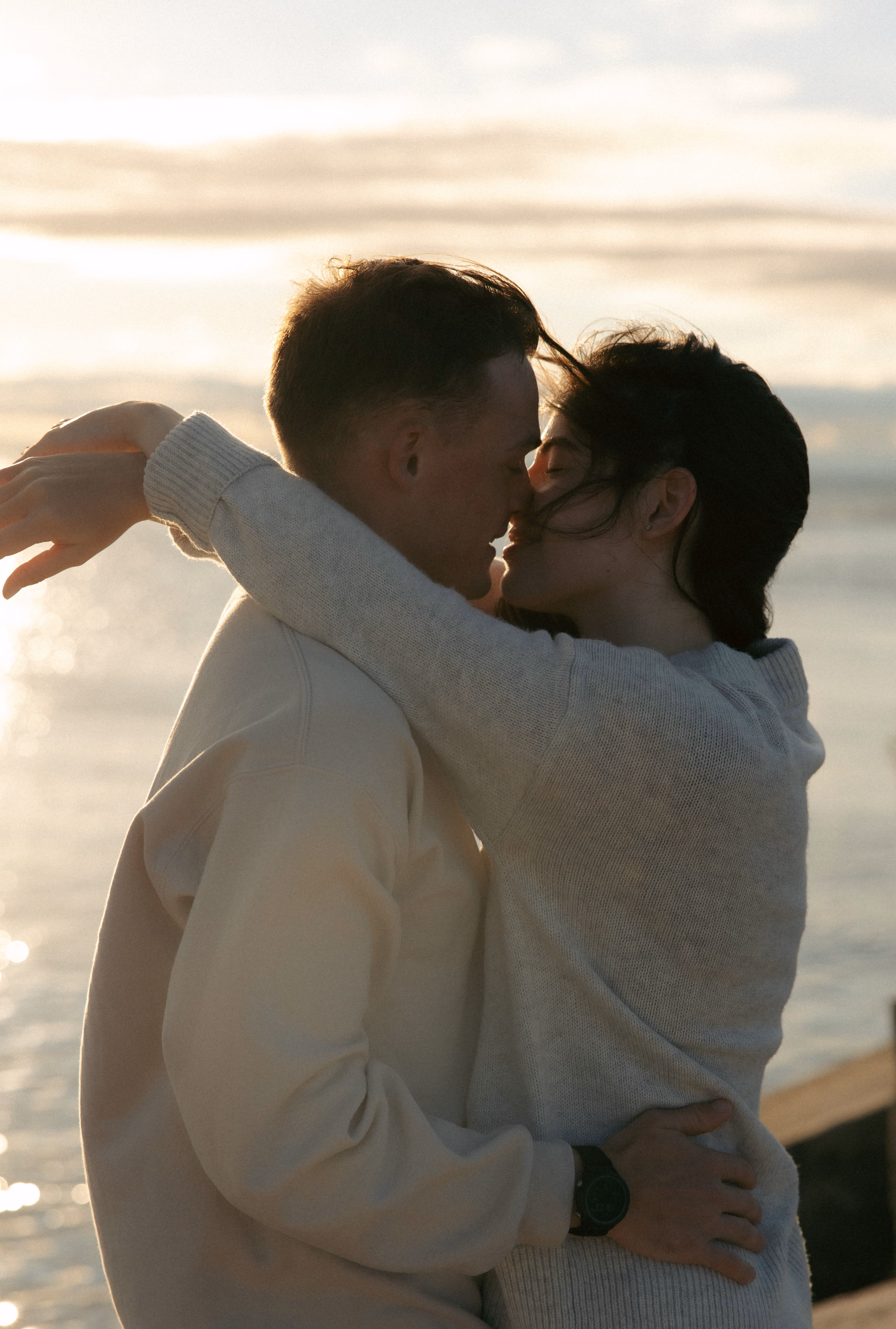 Couple embracing and about to kiss by water at sunset, wearing light sweaters.