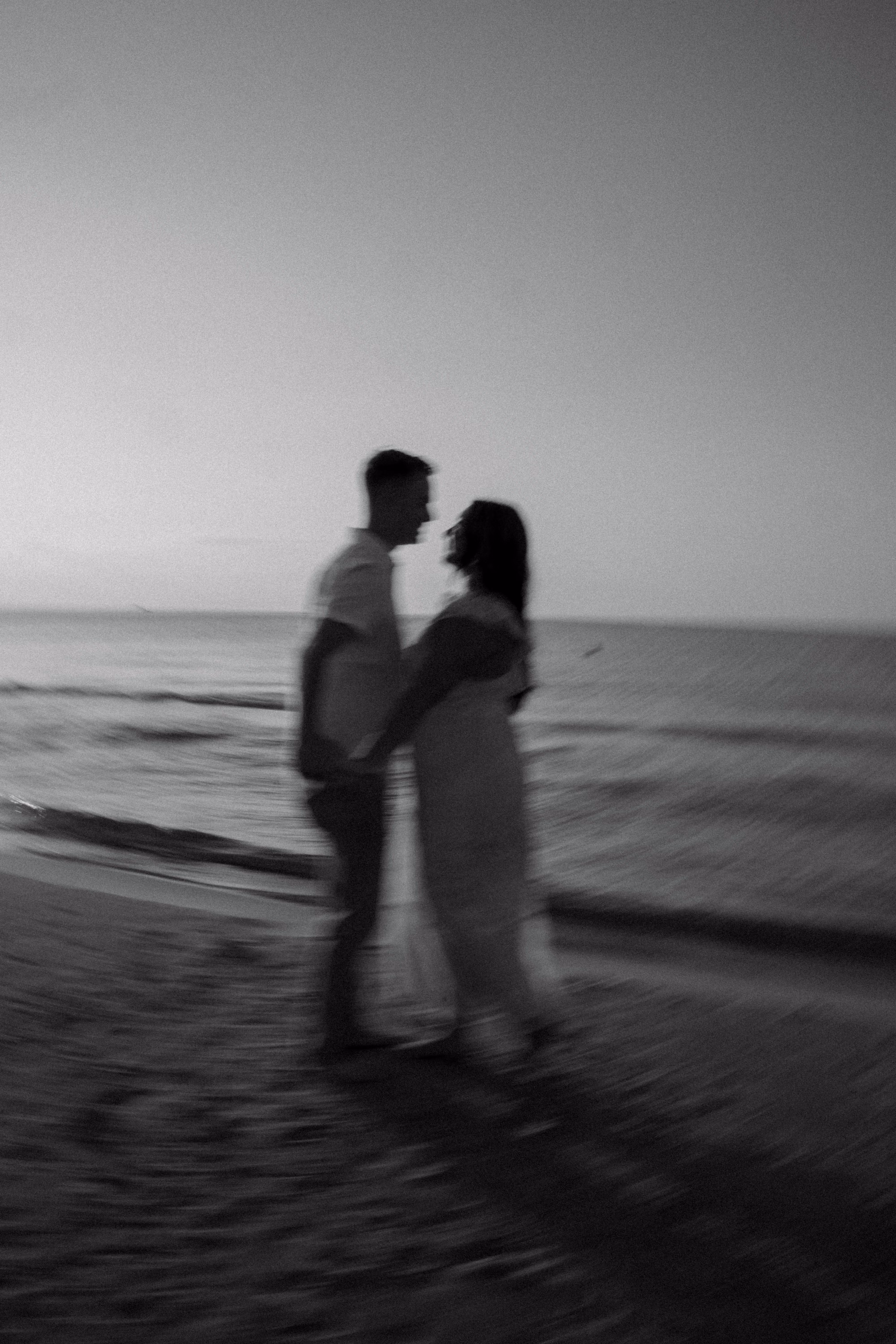 Blurred black and white photo of couple facing each other on a beach at sunset.
