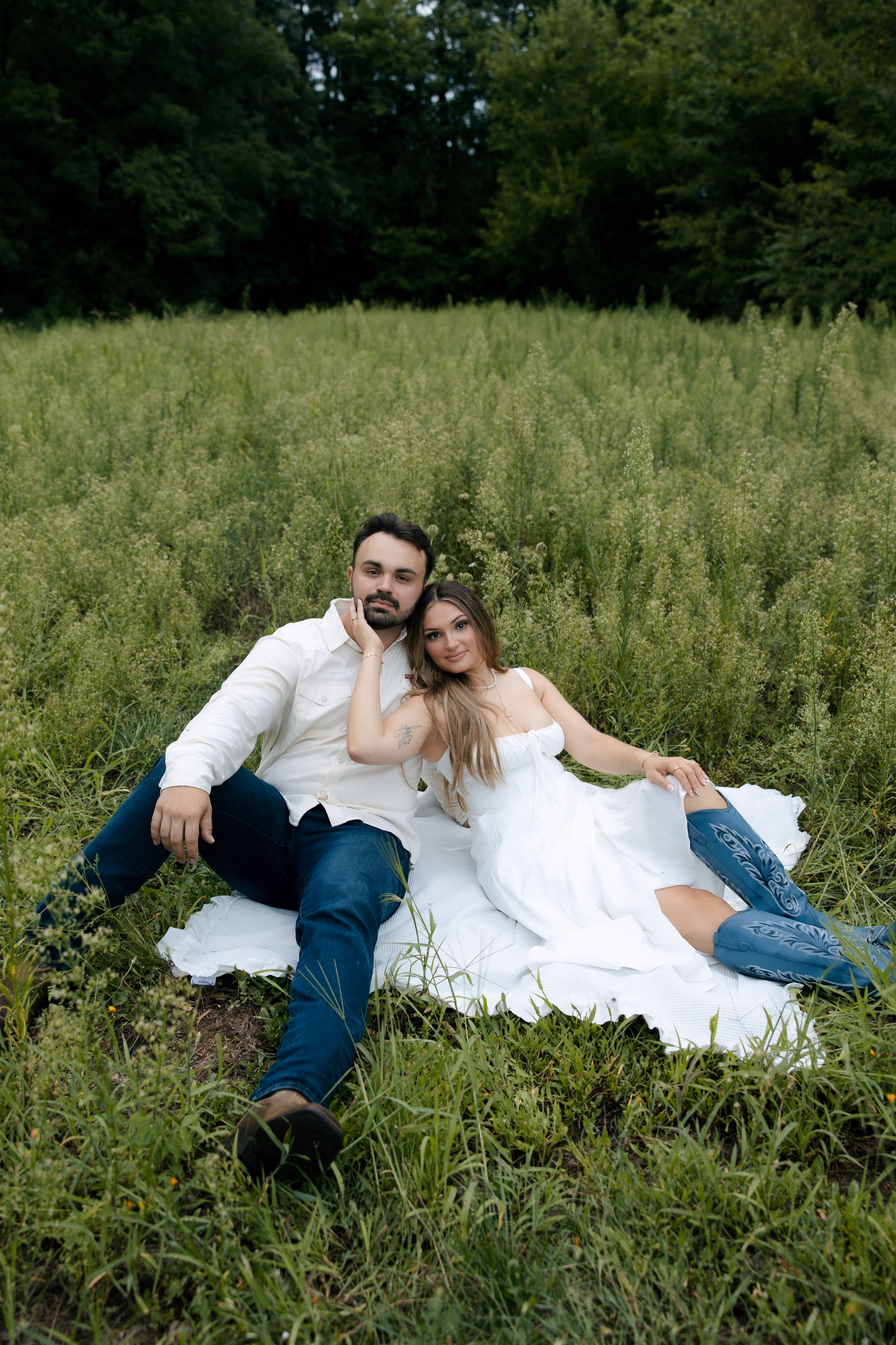 Couple sitting on a white blanket in a green field, the woman wearing a white dress and blue boots.