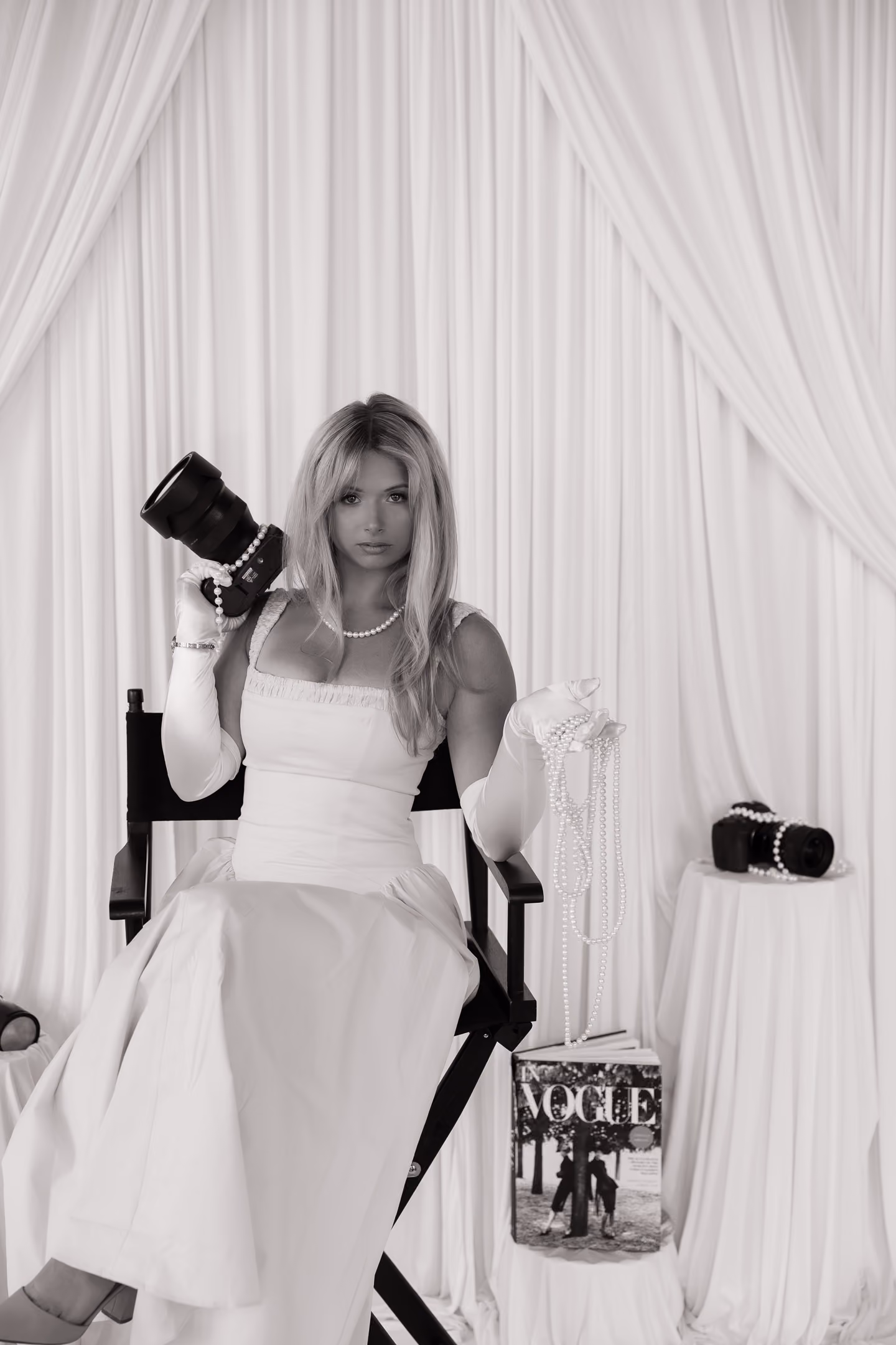 Reagan in white gown with pearls holding camera and pearls, sitting on director's chair in draped room.