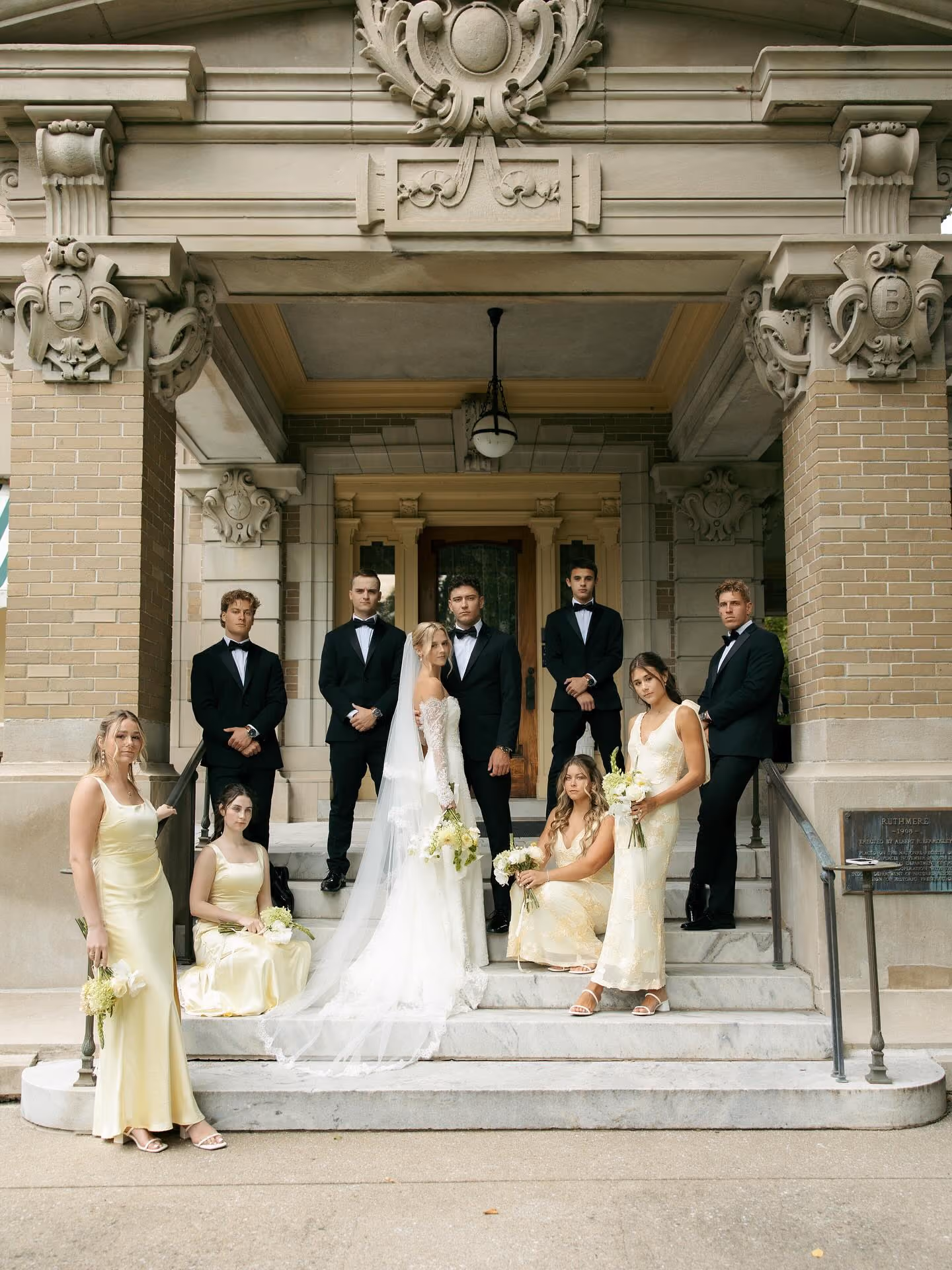 Bride holding white lilies as groom in white tuxedo kisses her temple outside a house.