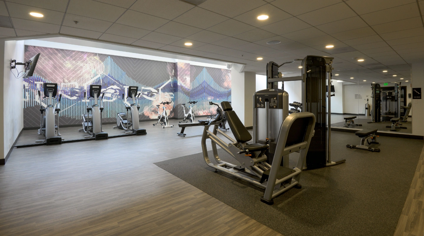 An empty, well-lit fitness center is equipped with a variety of exercise machines, including three elliptical trainers lined up against a wall with a colorful geometric mural, stationary bikes, a rowing machine, and a selection of weight machines and bench