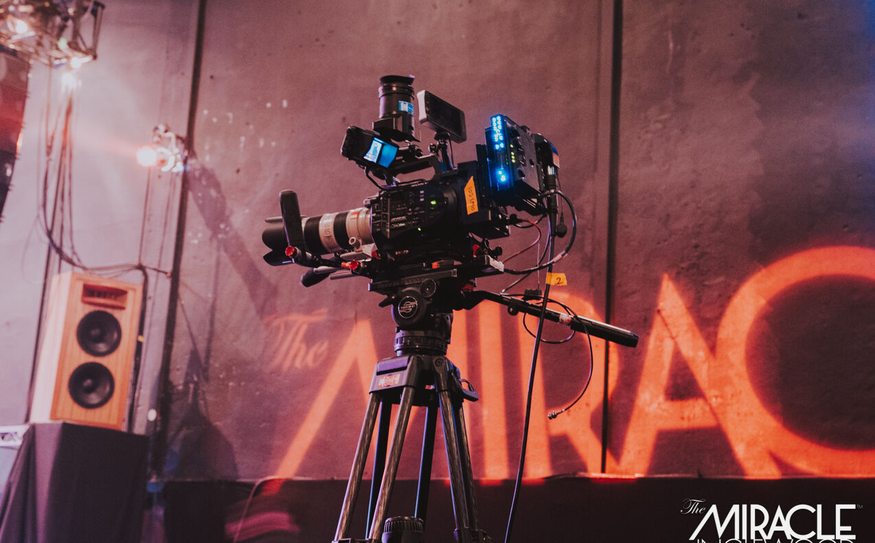 A cinema camera on sticks in front of the red Miracle wall