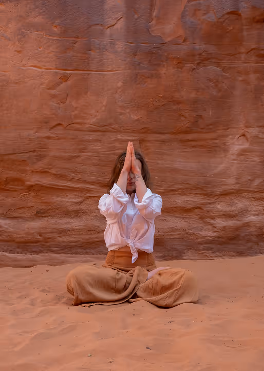 Woman sitting cross-legged on sand with hands pressed together in front of face near red rock wall.