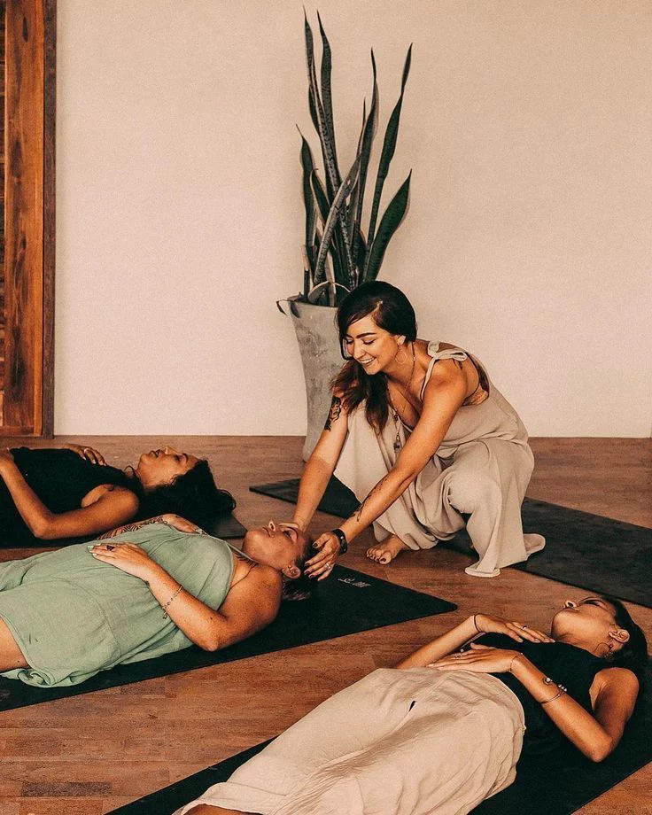 A woman guides three others lying on yoga mats in a calm, indoor meditation or relaxation session.