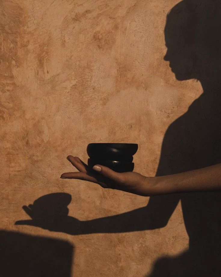Shadow of a person holding two black bowls against a textured brown wall.
