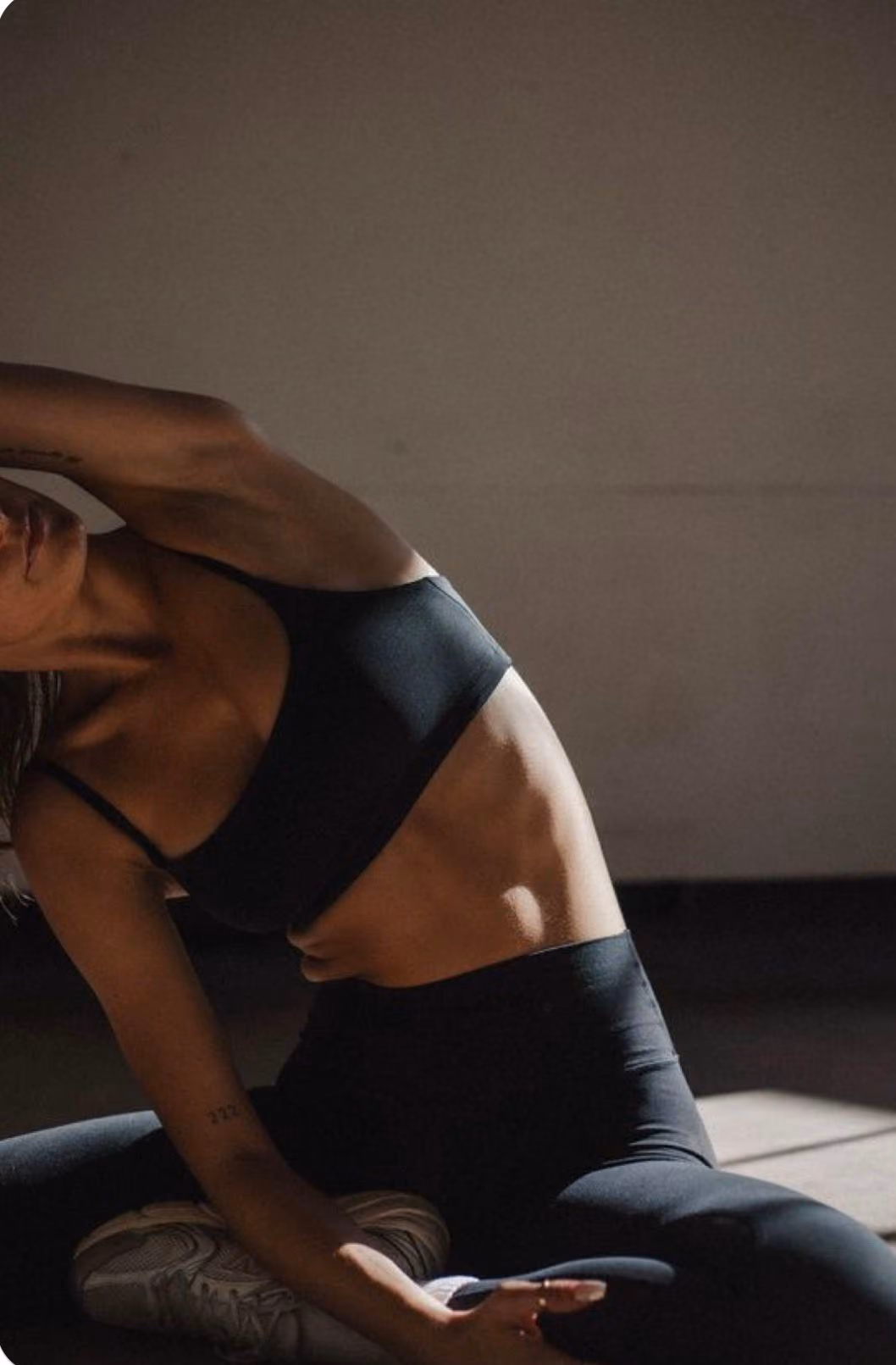 Woman in black sportswear stretching her side while sitting on the floor in a dimly lit room.