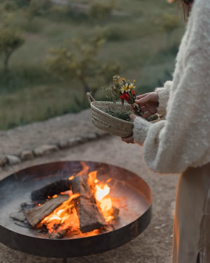 Person holding a woven basket with flowers near a lit outdoor fire pit with logs burning.
