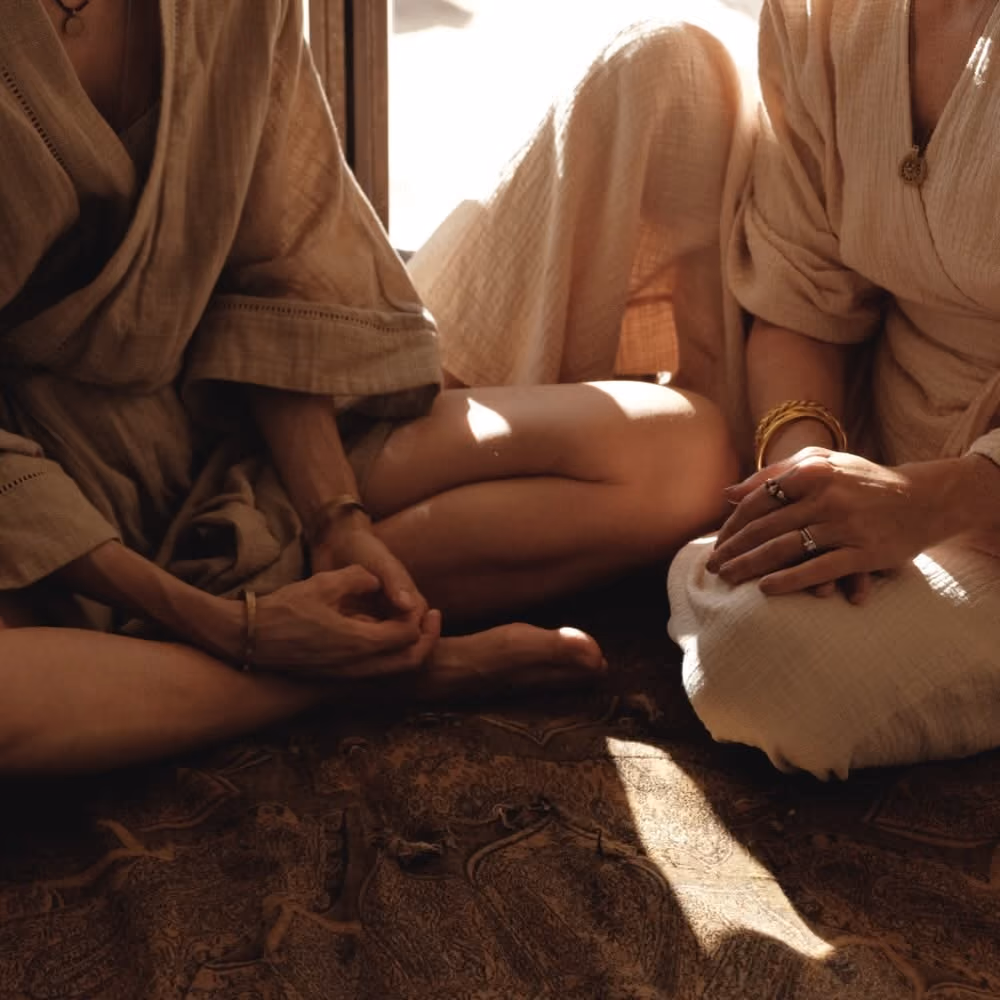 Two women sitting cross-legged on a patterned rug wearing beige robes with bracelets and rings.