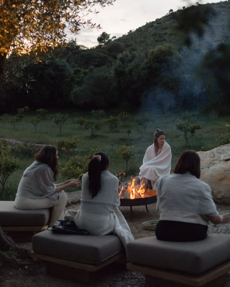 Four people wrapped in blankets sitting around a fire pit outdoors at dusk with trees and hills in background.