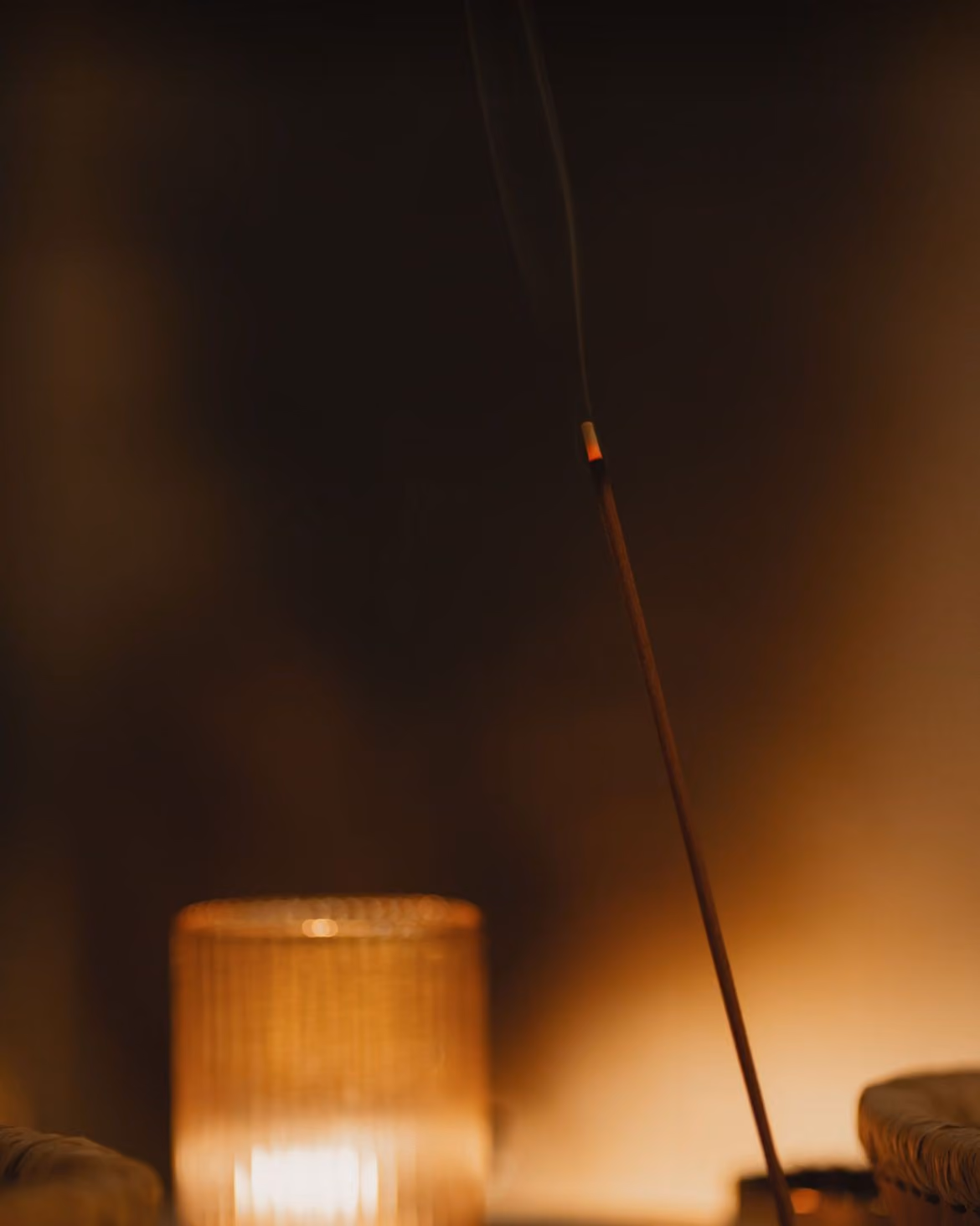 Burning incense stick emitting smoke with a lit candle blurred in the background.