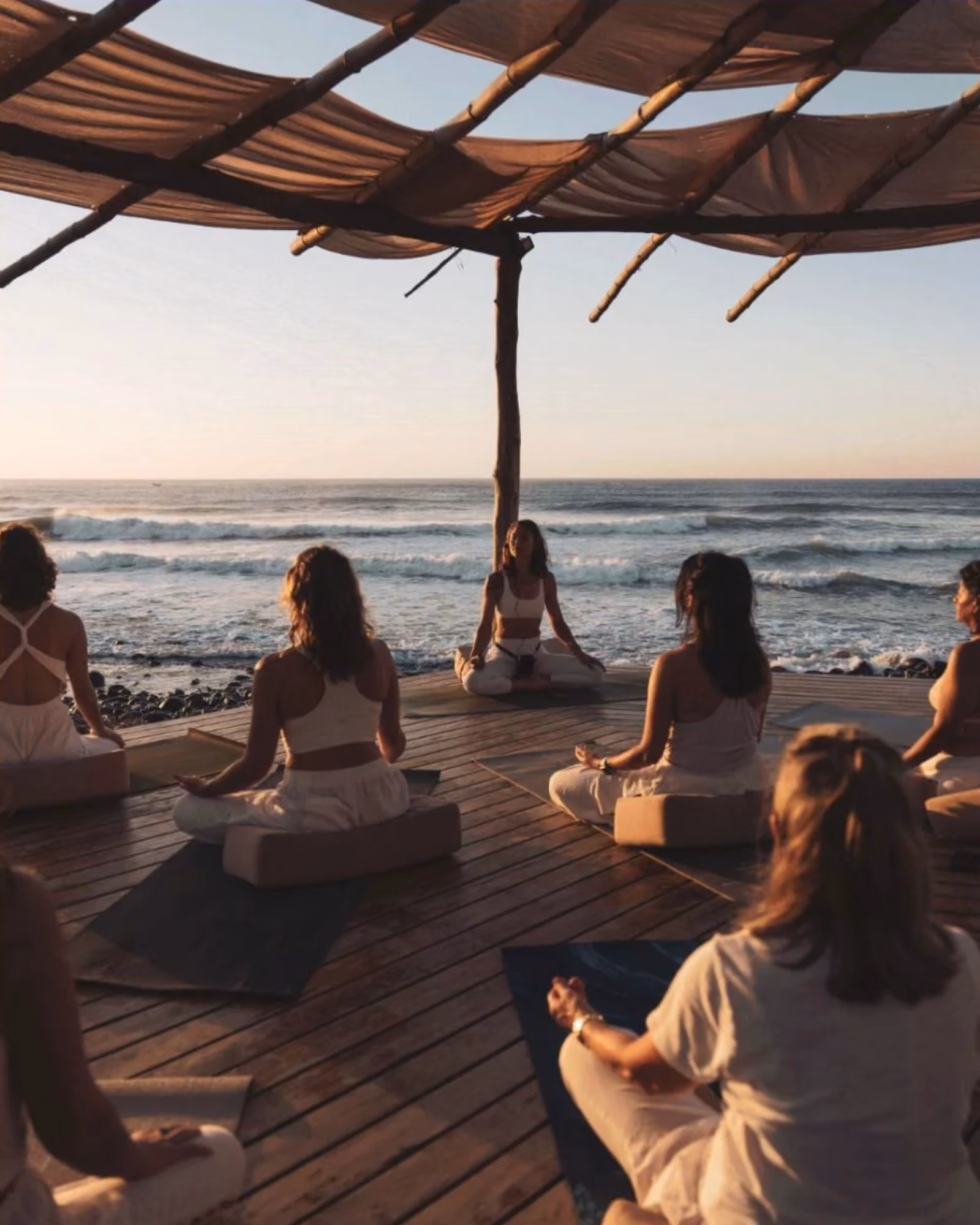 Group of people seated on cushions meditating on a wooden platform by the ocean at sunset.