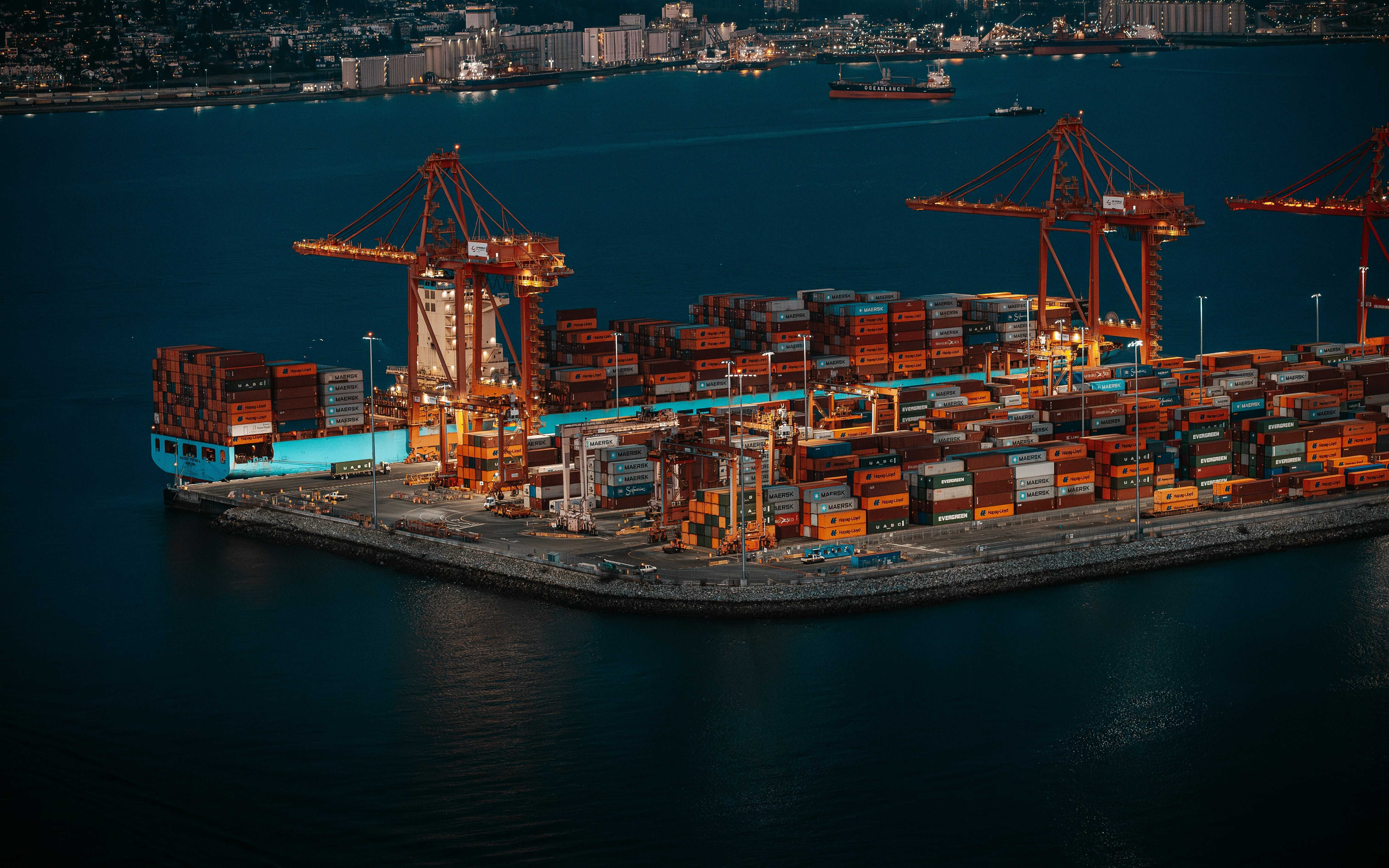Night view of a busy port terminal with cranes and stacked shipping containers on a dock.
