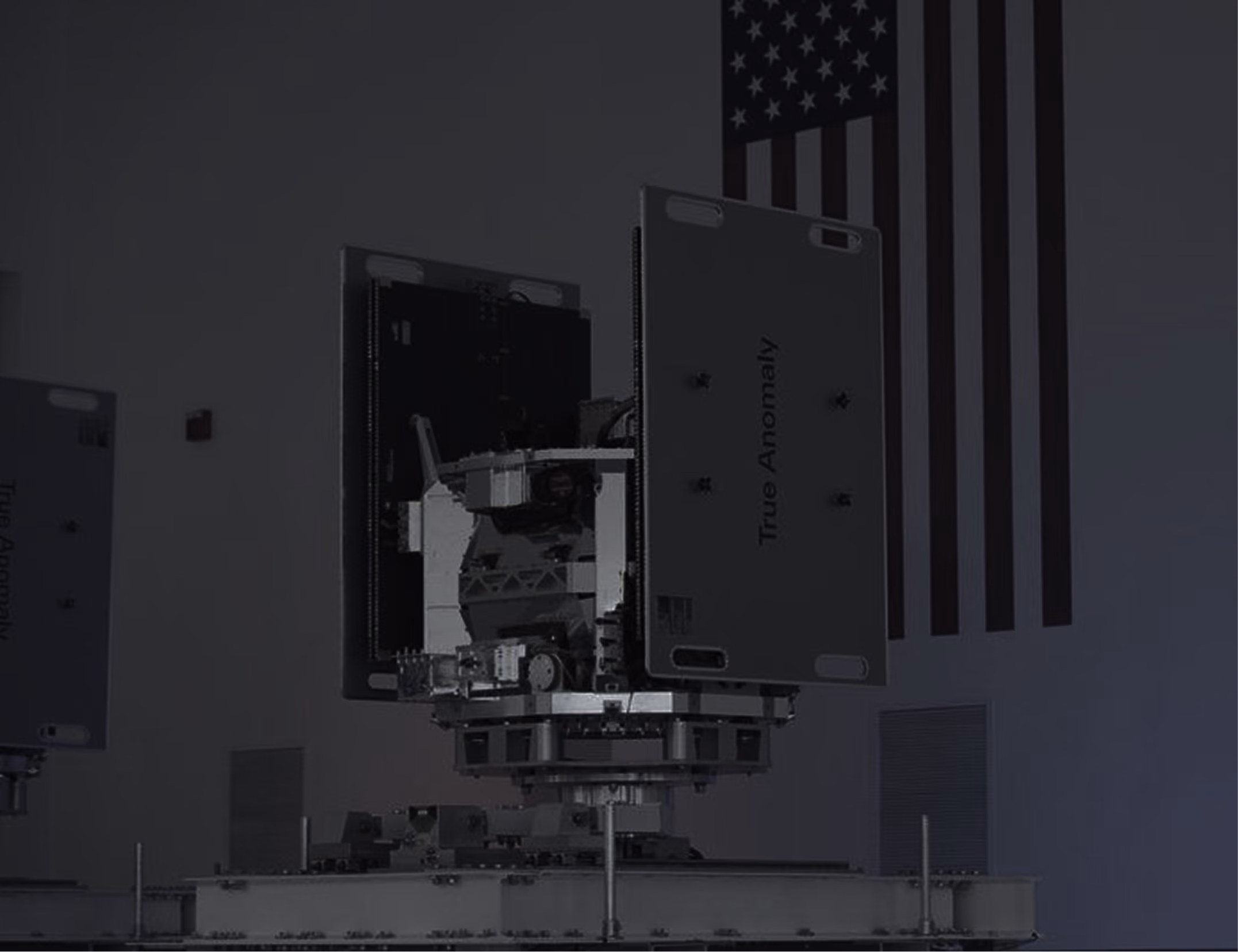 Satellite component labeled 'True Anomaly' mounted on a test rig with an American flag in the background.