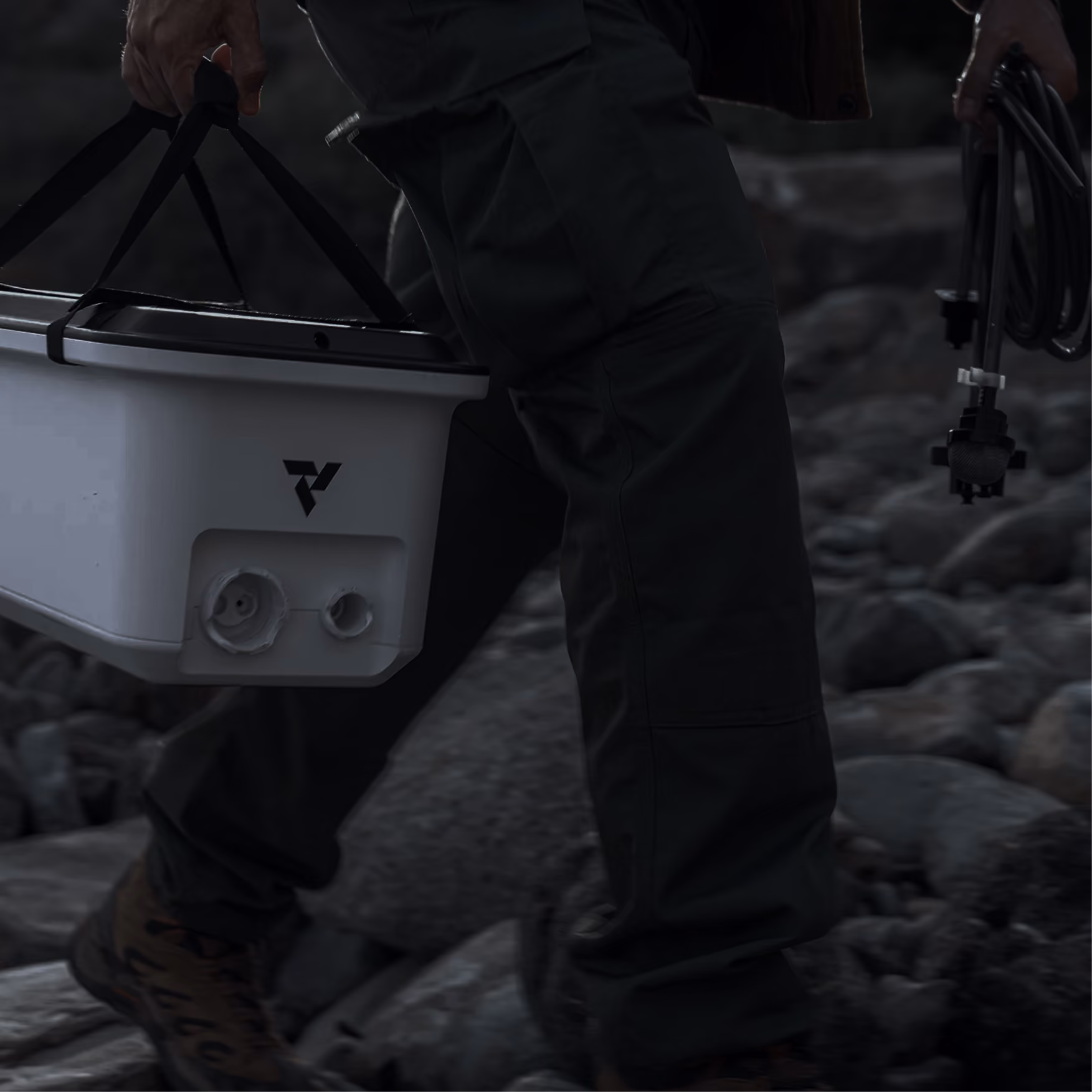 Person walking on rocky terrain carrying a white cooler and a coiled cable in each hand.