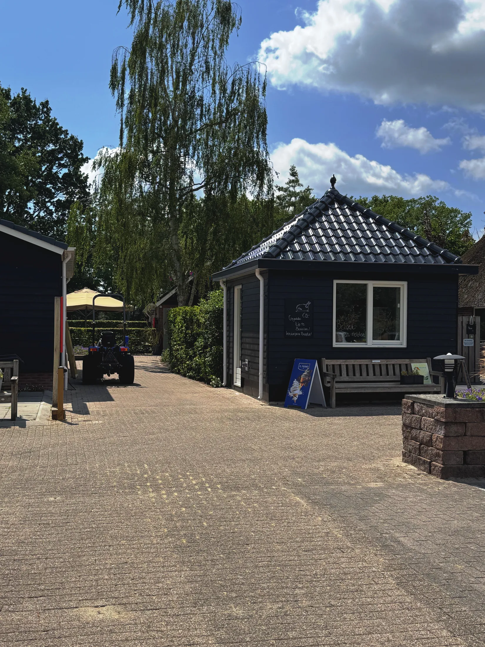 Oprit met boom, blauw gebouw en bank, blauwe lucht met wolken boven een camping.