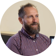 Man with a beard and short hair wearing a checkered shirt, smiling and looking to the side indoors.