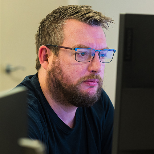 Man with glasses and a beard focused on a computer screen in an office setting.