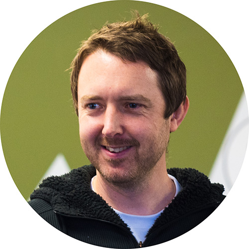 Smiling man with short brown hair and light beard wearing a black fleece jacket over a white shirt with an olive green background.