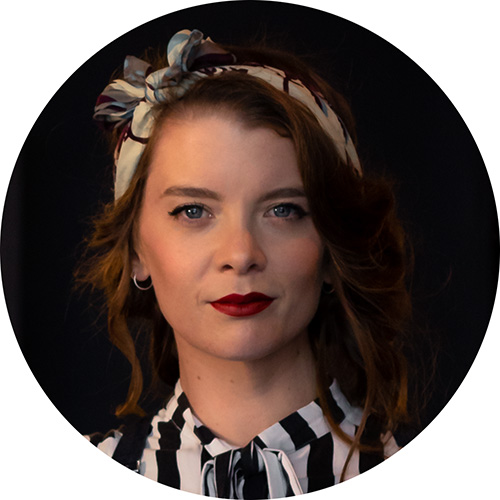 Portrait of a woman with curly brown hair wearing a floral headband and striped blouse against a dark background.