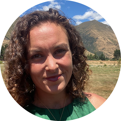 Smiling woman with curly hair wearing a green top outdoors with mountains and blue sky in the background.
