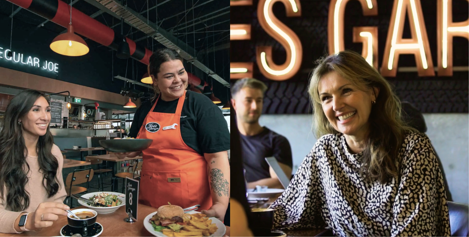 A smiling waitress in an orange apron serving food to a seated woman drinking coffee in a casual restaurant, with another woman smiling in the background.