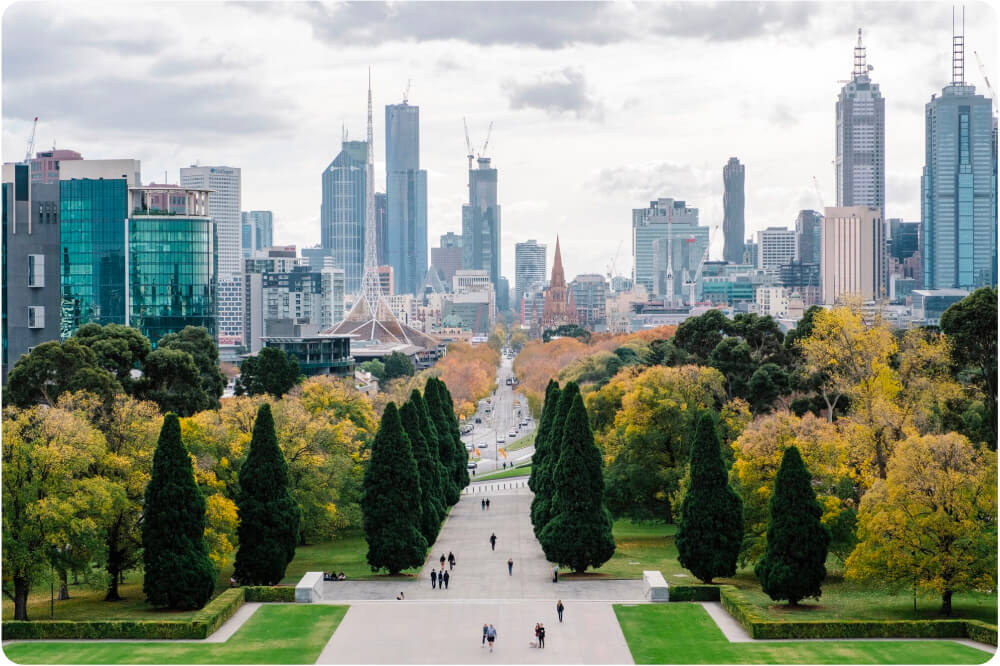 Wide view of Melbourne city skyline with tall buildings and a central park pathway lined with green and yellow trees in autumn.