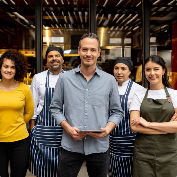 Smiling restaurant team of five people, including chefs in striped aprons and a man holding a tablet in front of a glass window.