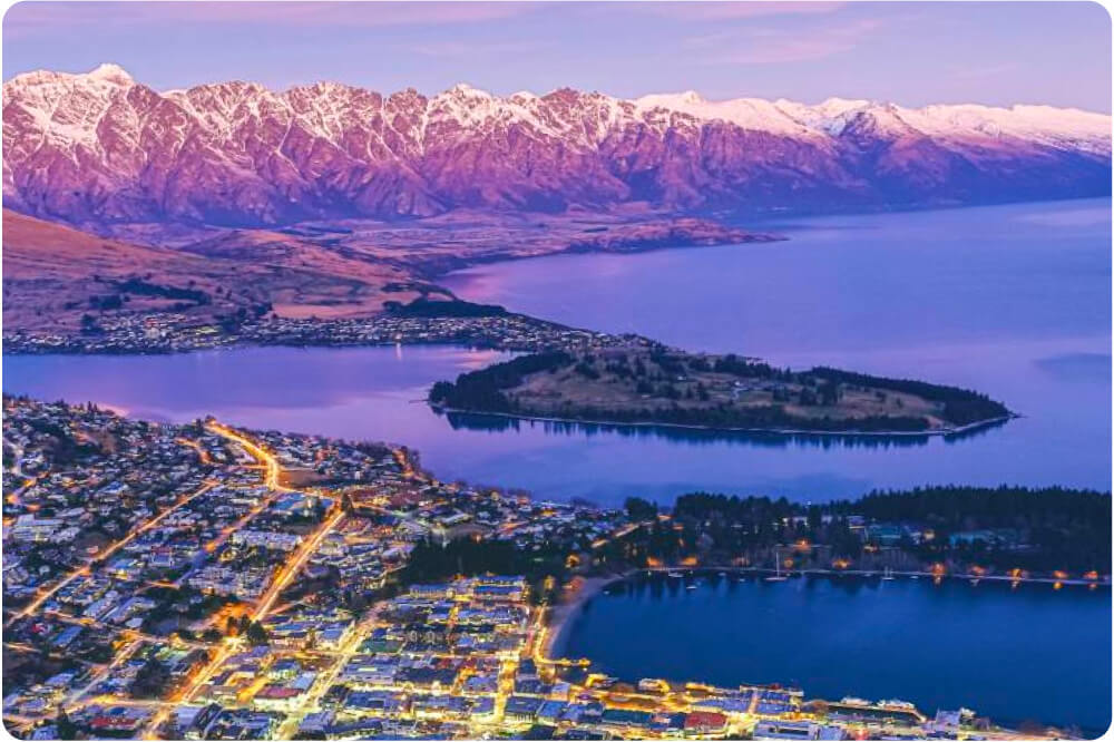 Aerial view of Queenstown at dusk showing illuminated streets, Lake Wakatipu, an island, and snow-capped mountains in the background.