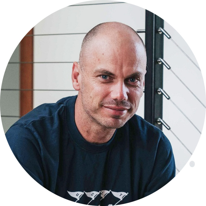 Portrait of a bald man with blue eyes wearing a dark shirt, sitting indoors near a railing with metal cables.