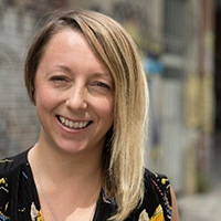 Smiling woman with blonde hair wearing a black floral top, standing outdoors in an urban setting.