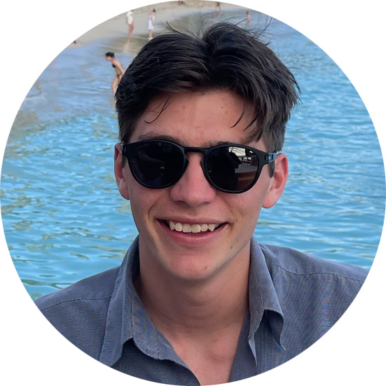 Young man with dark hair wearing black sunglasses and a gray shirt smiling near a body of water.