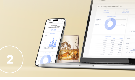 A smartphone displaying a sales dashboard with graphs sits next to a glass of whiskey on a counter beside an open laptop showing a similar dashboard.