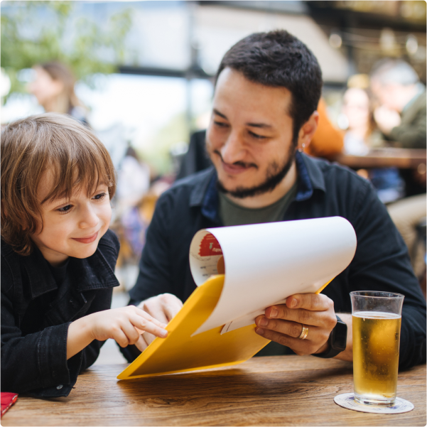 Man and young boy smiling while looking at a menu together at a wooden table with a glass of beer.