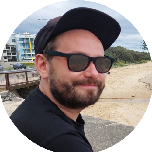 Bearded man wearing black sunglasses and a black cap sitting outdoors near a beach with buildings and trees in the background.