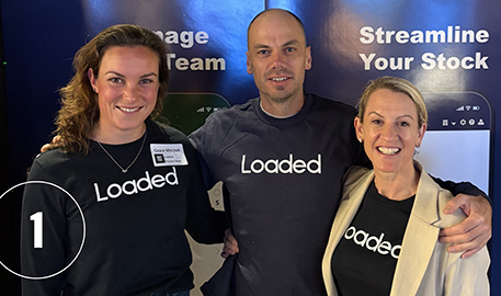 Three smiling team members wearing black Loaded shirts standing together in front of a blue background with white text.