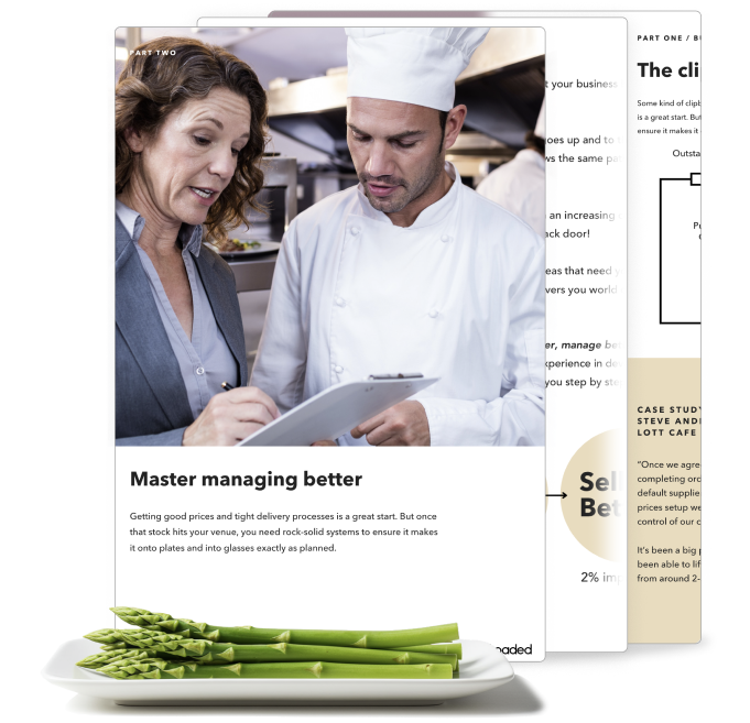Woman in business attire discussing notes on a clipboard with a chef in a kitchen, with a plate of fresh asparagus in front.