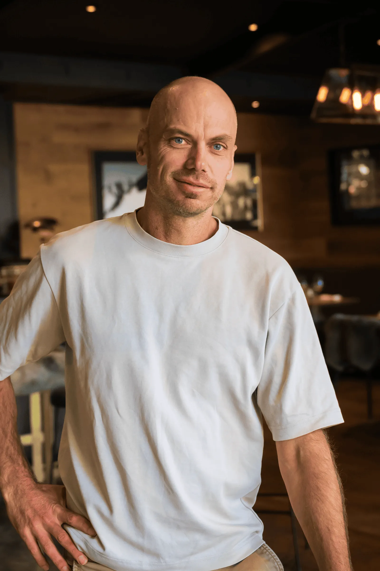 Bald man with blue eyes wearing a white t-shirt, standing indoors with a hand on his hip.