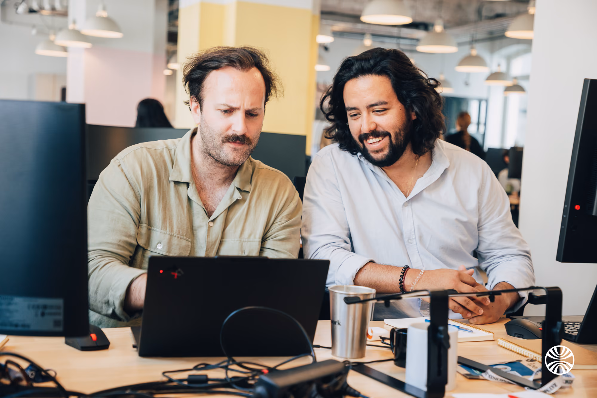 Two men working together on a laptop at a cluttered office desk with monitors and notebooks.