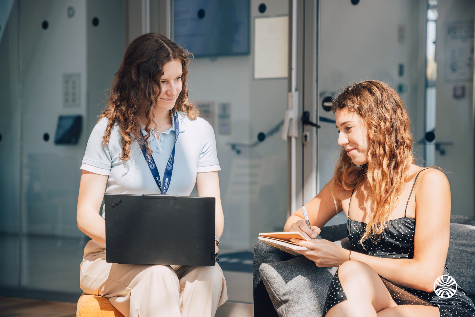 Two women working together indoors, one with a laptop, the other writing in a notebook.