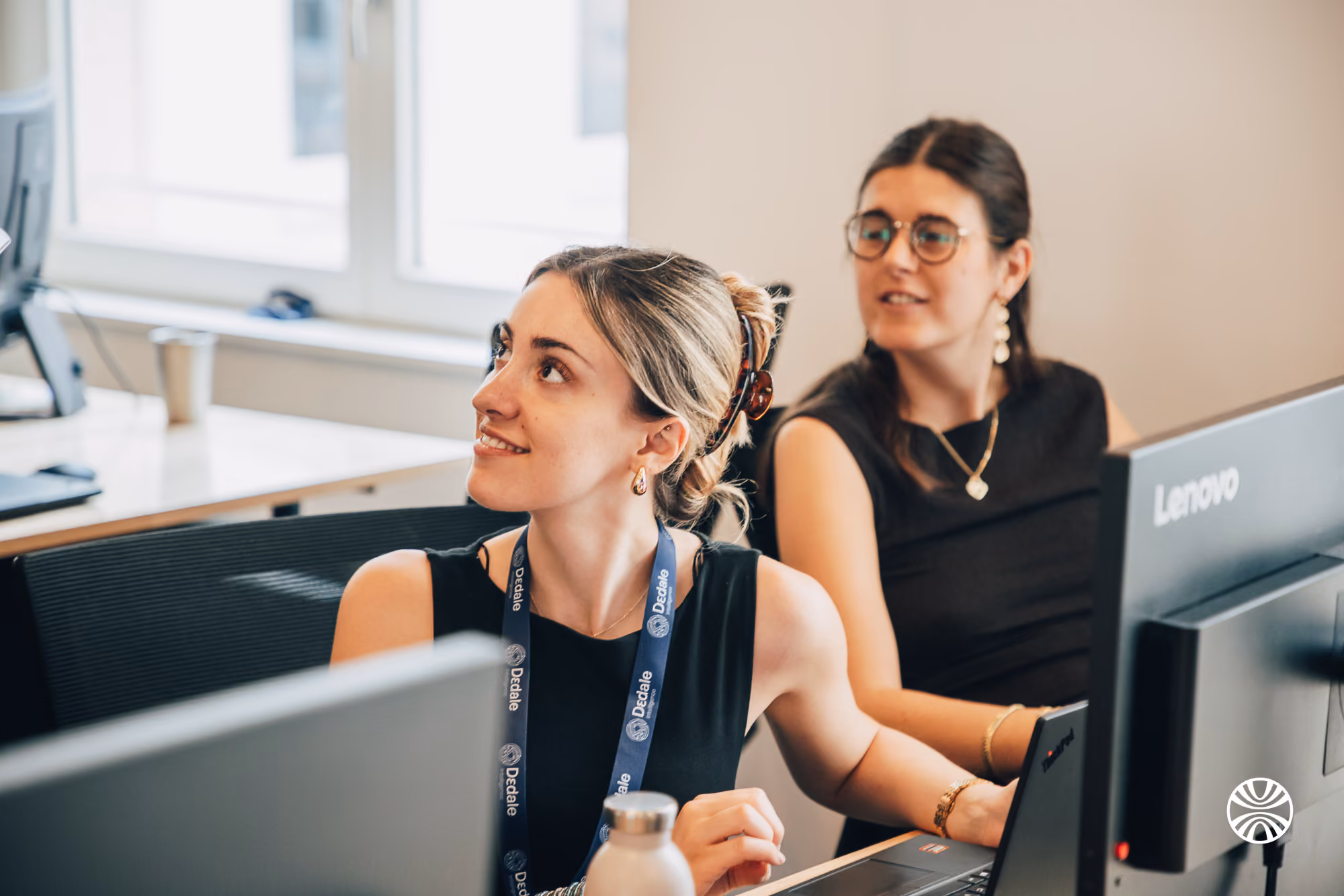 Two women at computers in an office, one looking up and the other looking at her screen.