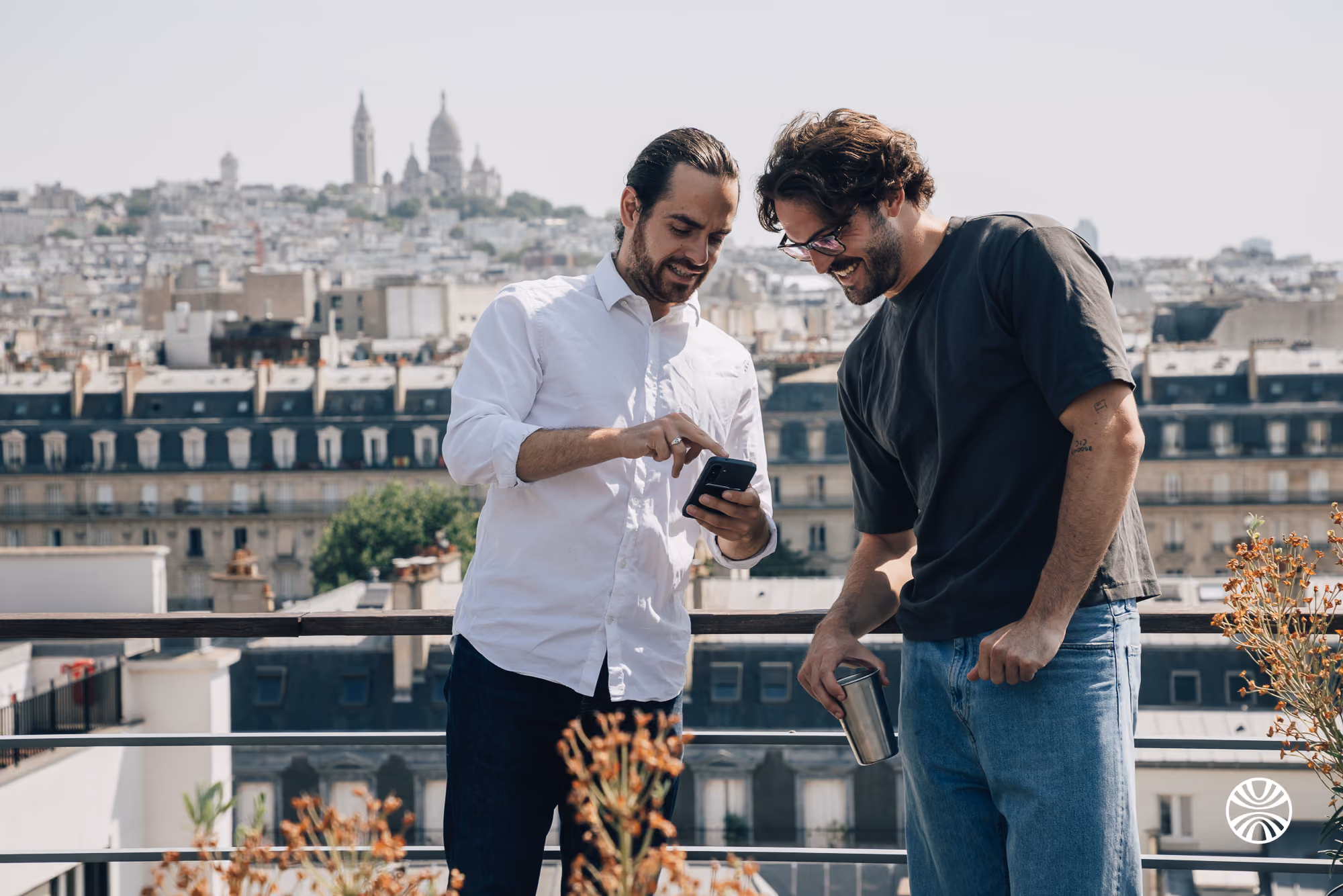Two men on a rooftop terrace looking at a phone, with city buildings and a cathedral in the background.