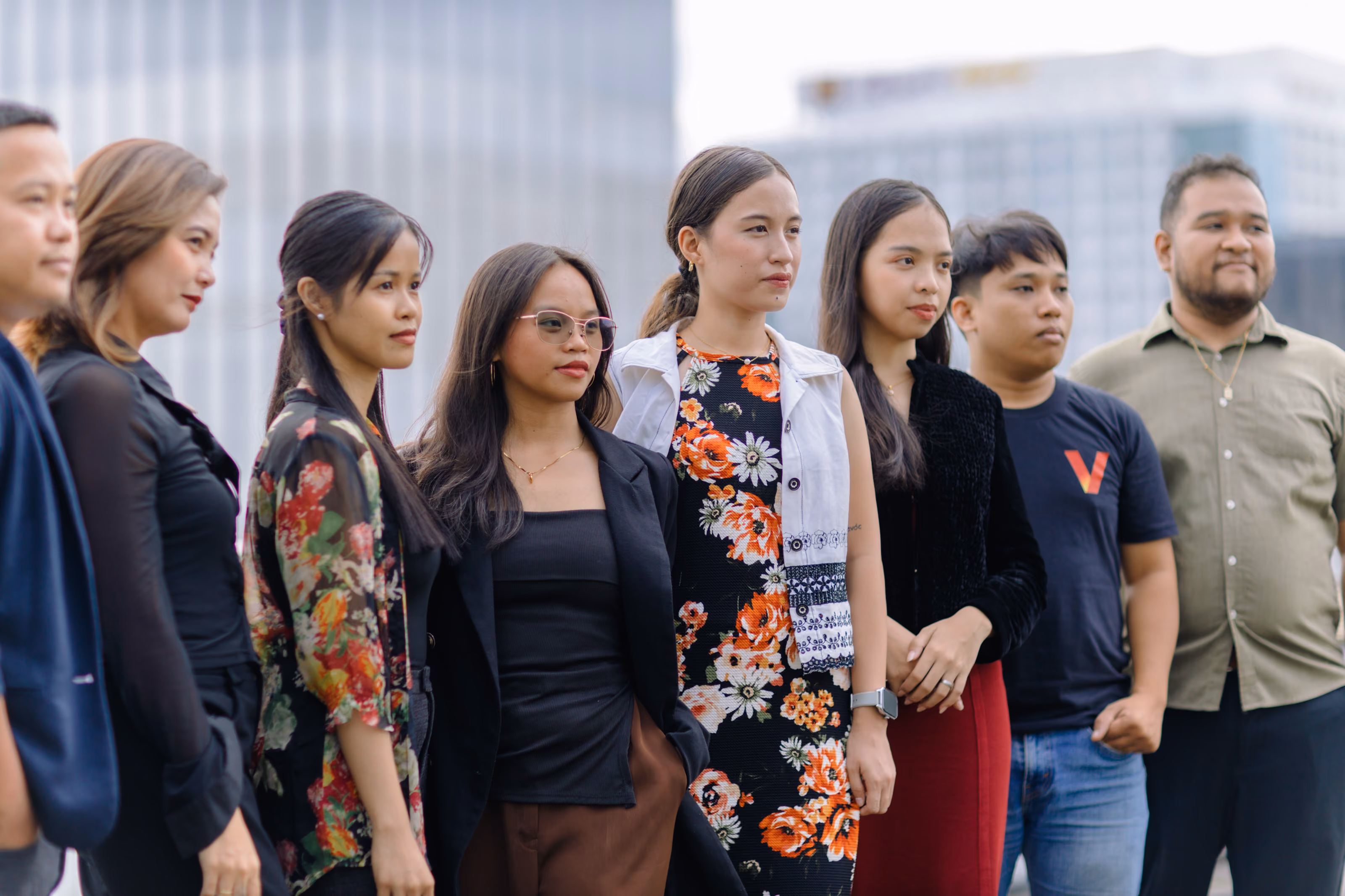 Seven diverse adults standing in a row outdoors with buildings blurred in the background.