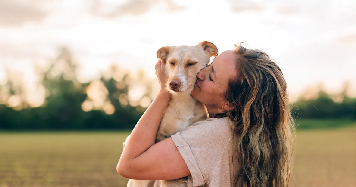 Woman with long hair hugging and kissing a light brown dog outdoors in a field during sunset.