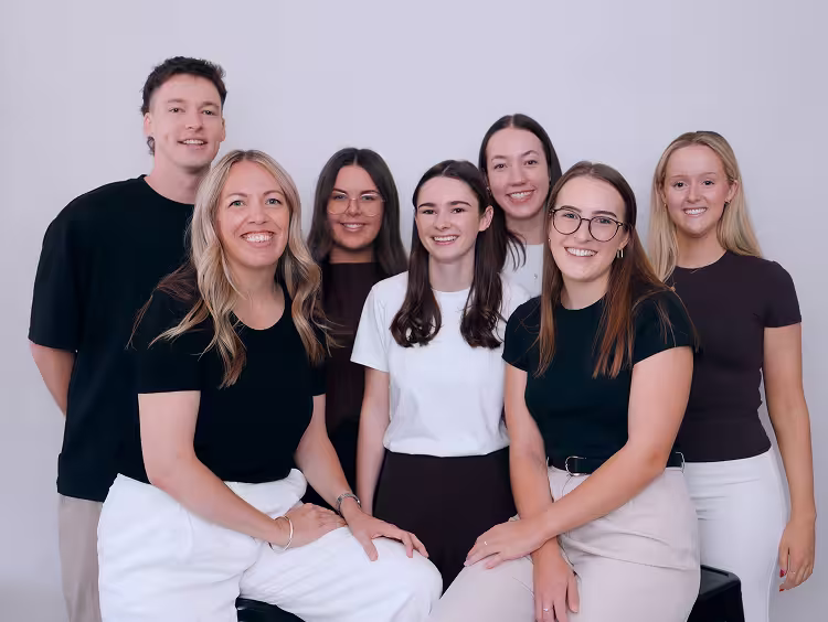 Group of seven young adults smiling and posing against a plain light-colored background.