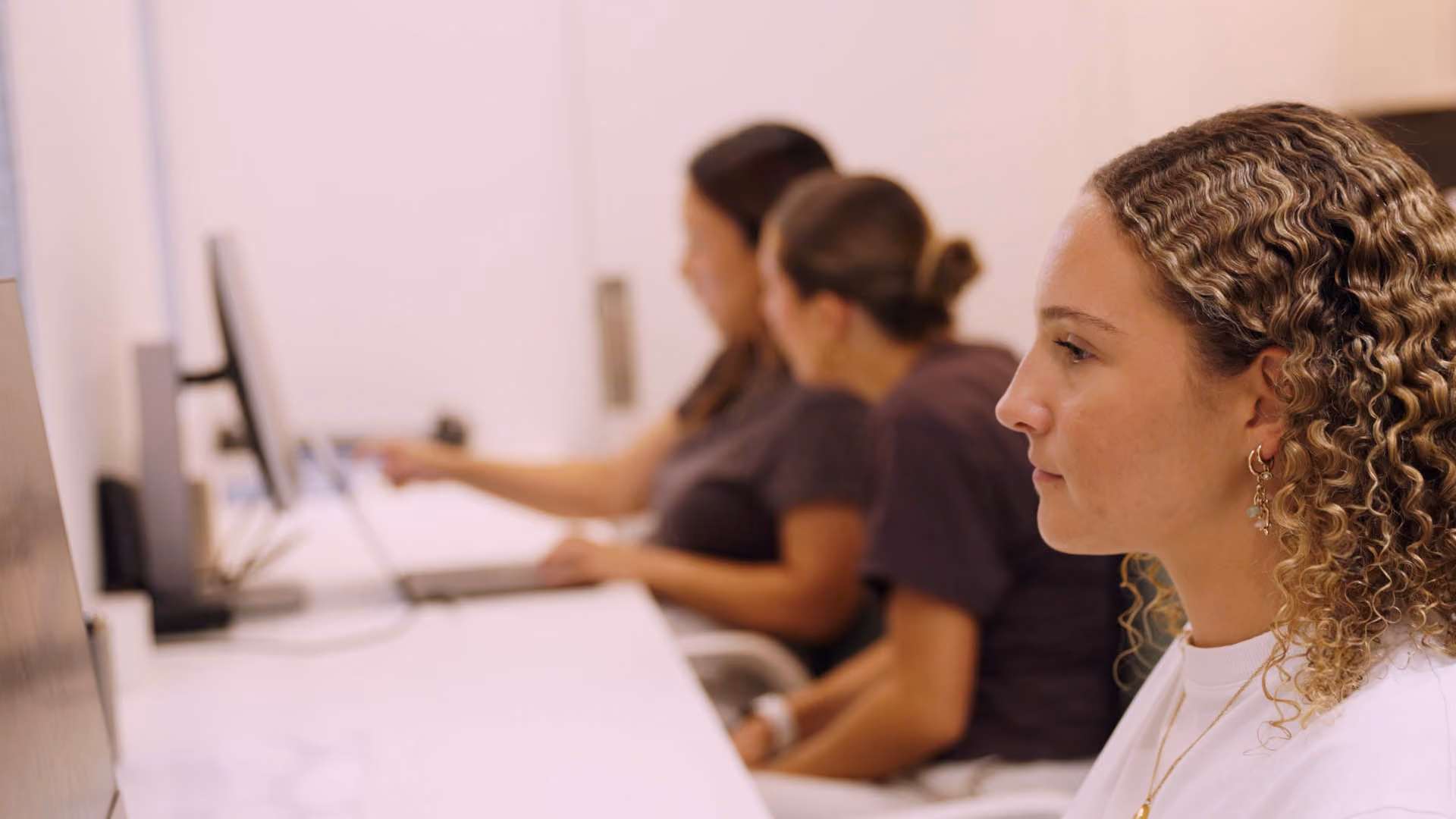 Three women sitting at desks working on desktop computers in a bright office space.