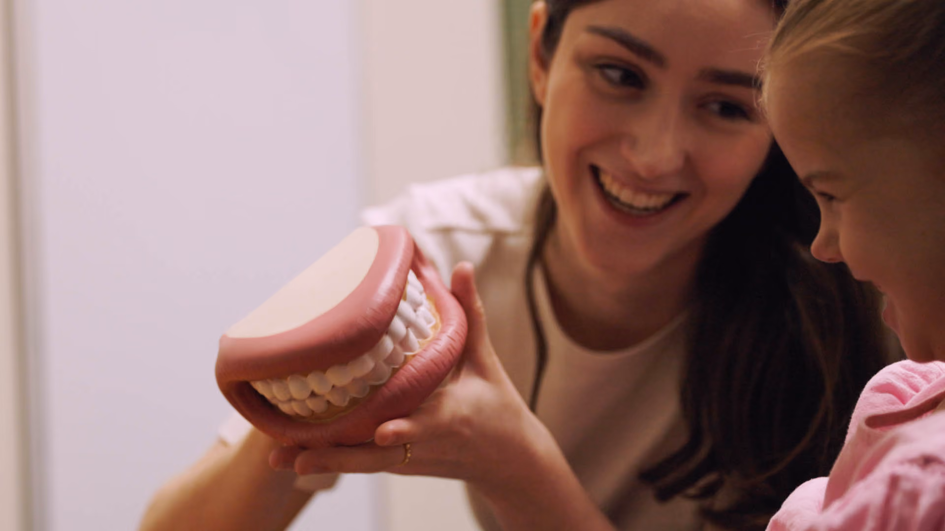 Woman smiling and showing a dental model of teeth to a young girl who is also smiling.